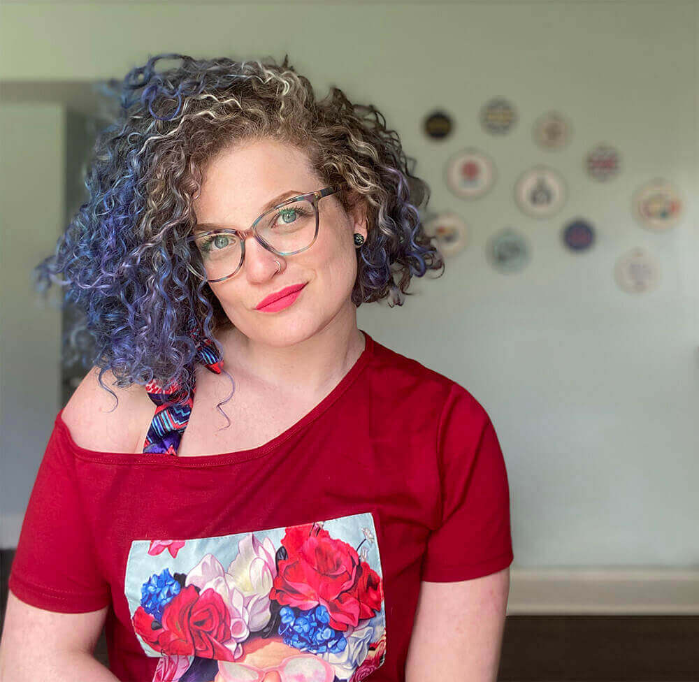 Portrait of Sex Coach & Educator Rachael Rose. A white woman with blue and purple curly hair in a red shirt softly smiling at the camera in front of a wall of embroidery art.