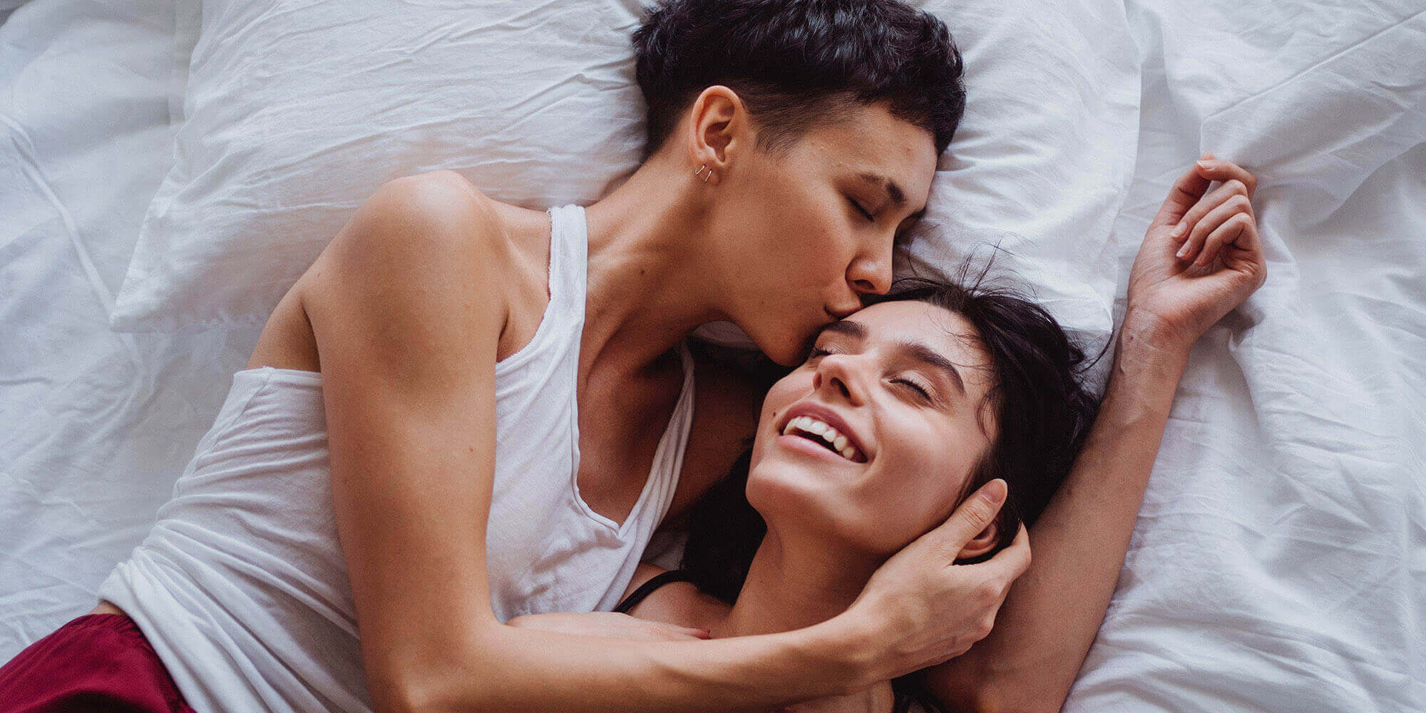 Two women cuddling in bed, and one is kissing the other's forehead